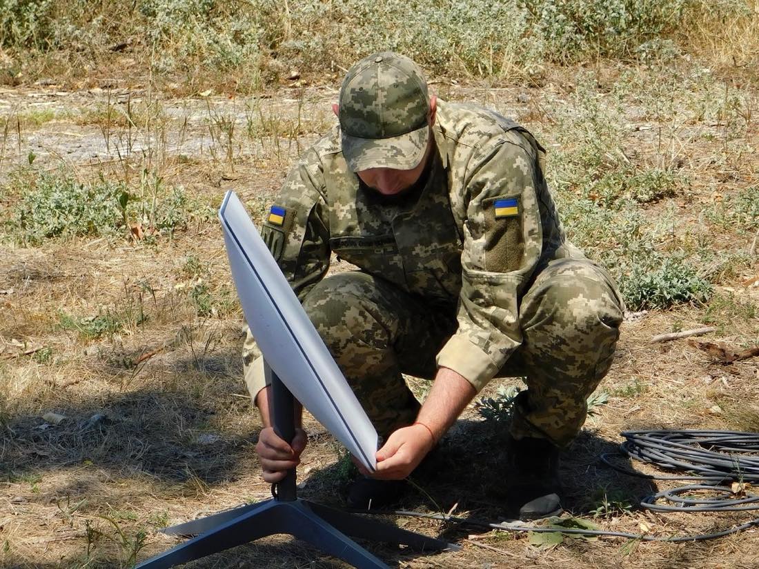 A Ukrainian soldier in camouflage kneels to set up a white Starlink satellite dish on a stand in a dry field.